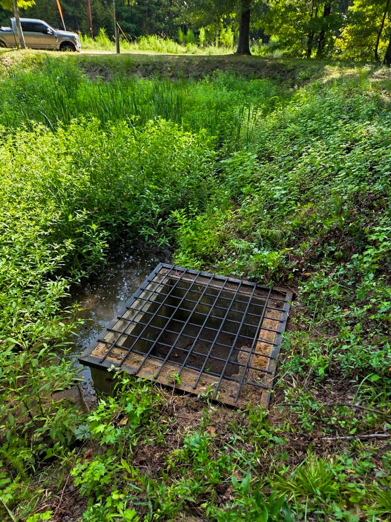 Metal debris screen over a concrete drainage structure surrounded by vegetation and shallow water.