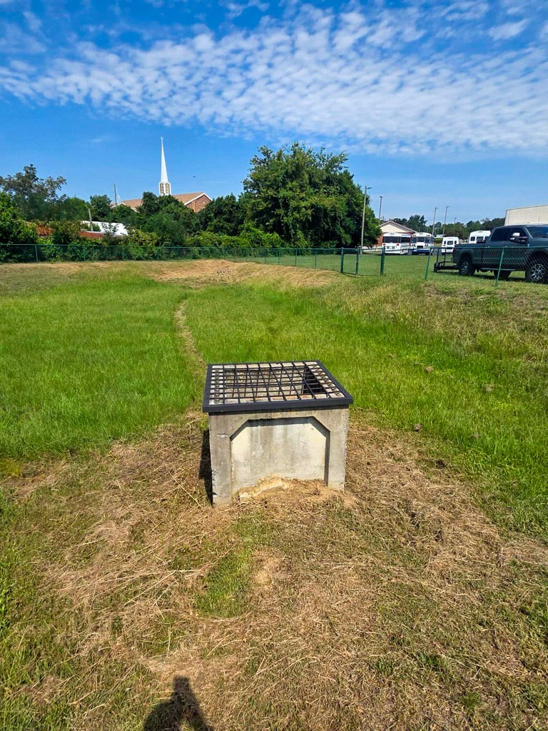 Square metal trash rack covering a concrete outlet structure in a grassy stormwater basin.